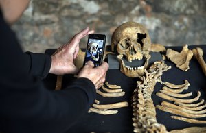 In this Wednesday, March 26, 2014 photo, a member of the media photographs the scull of one of the skeletons found by construction workers under central London's Charterhouse Square. The 25 skeletons were uncovered last year during work on Crossrail, a new rail line that's boring 13 miles (21 kilometers) of tunnels under the heart of the city. Archaeologists immediately suspected the bones came from a cemetery for victims of the bubonic plague that ravaged Europe in the 14th century. The Black Death, as the plauge was called, is thought to have killed at least 75 million people, including more than half of Britain's population. (AP Photo/Lefteris Pitarakis)
